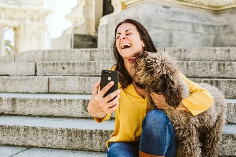 a woman laughing as her dog licks her and she takes a selfie
